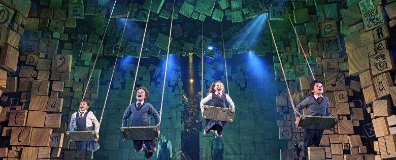 Matilda on stage. 4 children are on their bellies on swings and in their school uniform