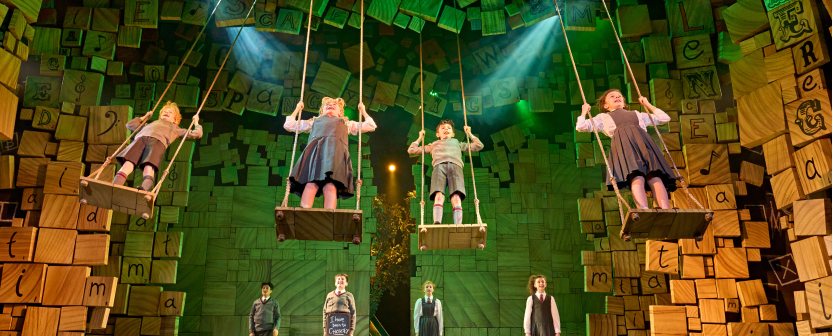 Children's cast of Matilda the Musical perform on stage. 4 are on swings, 4 are pictured at the back of the stage. Alphabet decor decks the edges of the stage.