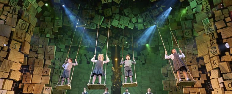 Children's cast of Matilda the Musical perform on stage. 4 are on swings, 4 are pictured at the back of the stage. Alphabet decor decks the edges of the stage.