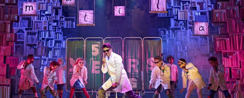 Cast of Matilda the Musical performing on stage. They wear lab coats and stethoscopes.