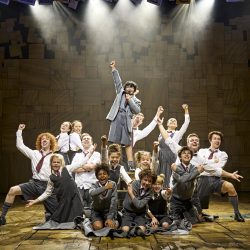 Cast of Matilda on stage. A group of children in their school uniform in the centre of the stage. Many lift their arms in triumph.
