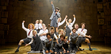 Full company of Matilda The Musical during the end of Revolting Children. They are posed with their arms in the air.