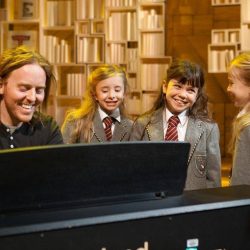 Tim Minchin, creator of Matilda The Musical, plays the piano with 3 Matilda's next to it smiling and sining.